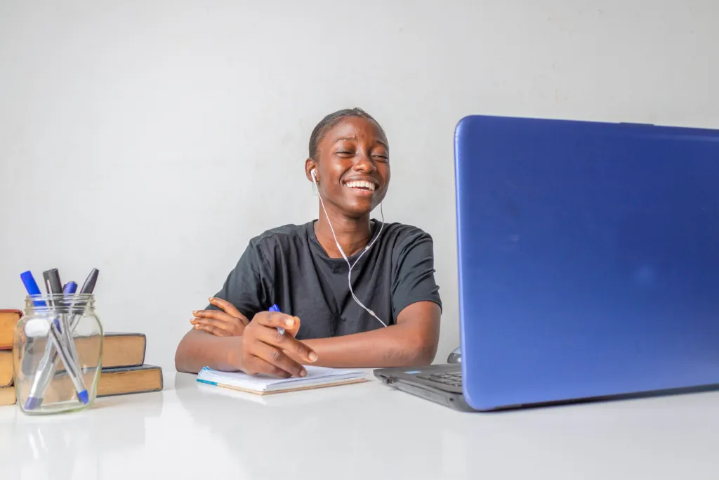 A young student in a gray shirt smiles while sitting at a desk and looking at a laptop.