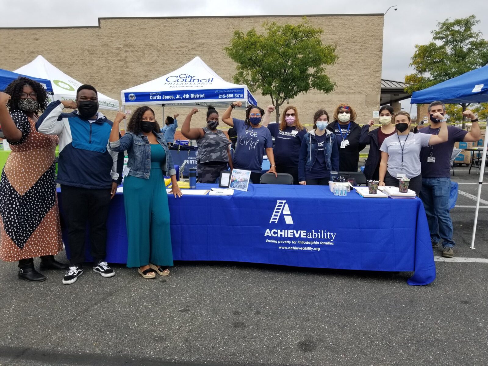 A group of masked people, many of whom are flexing an arm, stand behind and next to a table with a blue tablecloth that reads 