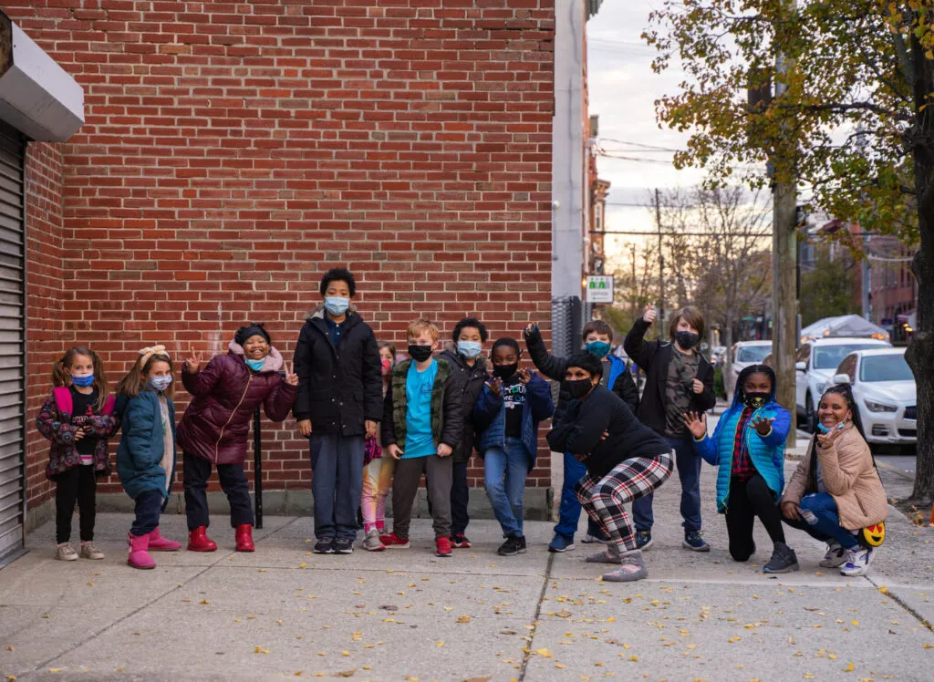 A group of 12 young children in winter coats and face masks pose with an adult in a face mask, black sweater, and plaid pants, in front of a brick building