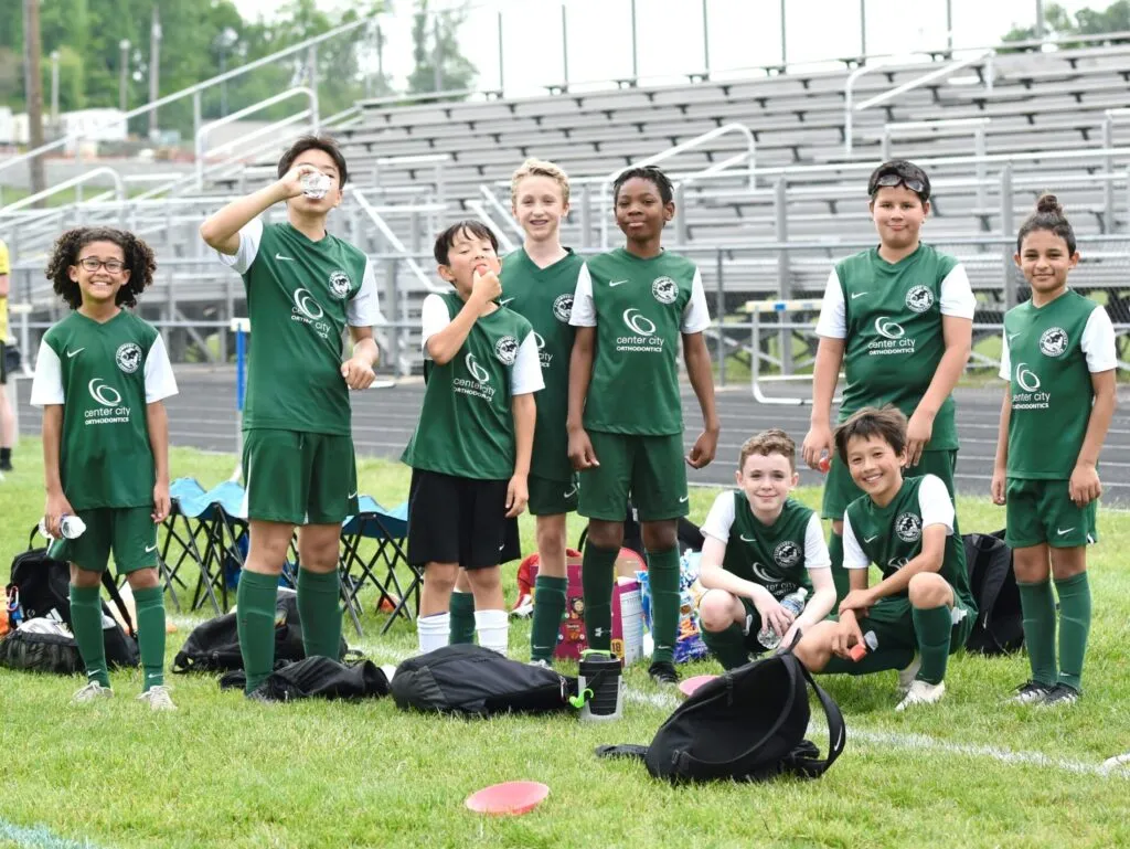 A group of children dressed in green soccer uniforms pose for a photo on a field in front of bleachers.