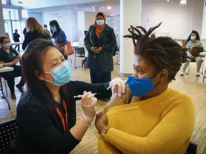 A person with black hair wearing a black sweater and face mask administers a vaccine to a woman with dark brown hair in a yellow sweater wearing a face mask.