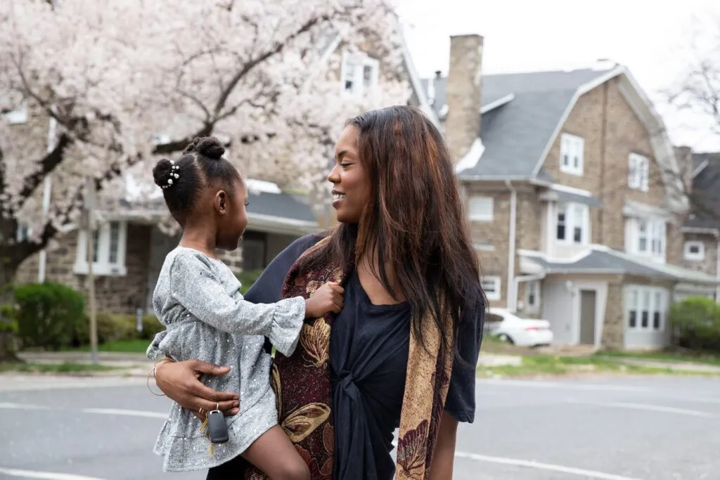 A person with long brown hair wearing a black dress and multicolor scarf stands in front of a row of homes and a flowering tree while holding a young child with dark hair wearing a gray dress