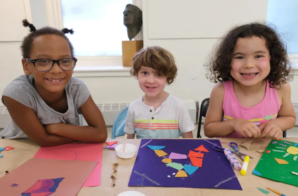 Two children are seated at a table, smiling and engaging in an arts and crafts activity. The child on the left is wearing glasses and a gray shirt, leaning on the table with arms crossed. The child on the right, with curly hair and a white shirt with colorful stripes, is smiling directly at the camera. In front of them are various colorful craft materials, including construction paper, scissors, and glue.