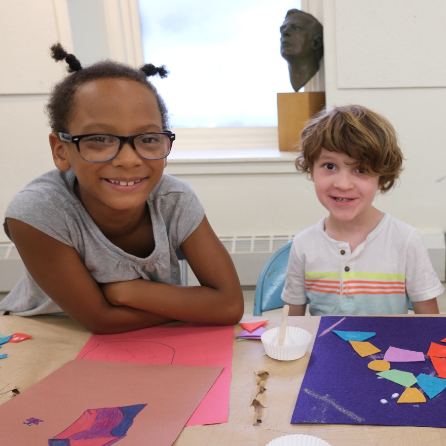 Two children are seated at a table, smiling and engaging in an arts and crafts activity. The child on the left is wearing glasses and a gray shirt, leaning on the table with arms crossed. The child on the right, with curly hair and a white shirt with colorful stripes, is smiling directly at the camera. In front of them are various colorful craft materials, including construction paper, scissors, and glue.