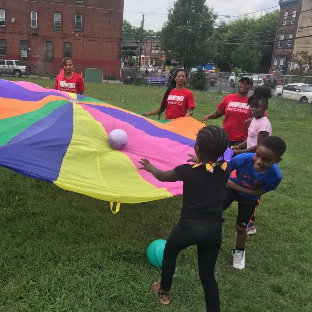 A group of children and adults are playing with a colorful parachute outdoors in a grassy area. The children are bouncing a ball on the parachute while the adults, wearing red shirts, guide the activity, with a backdrop of urban buildings and trees.
