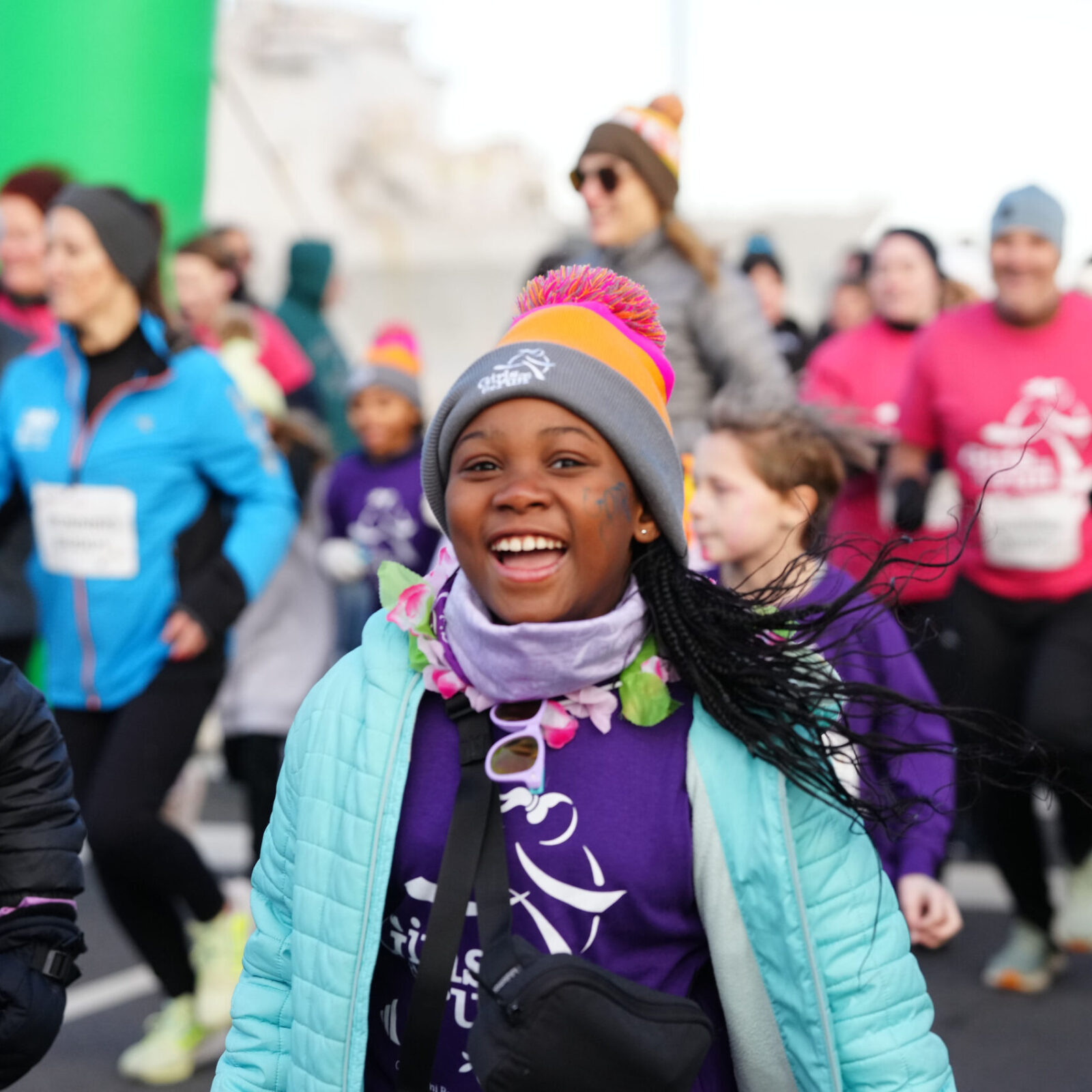 A child smiles widely while wearing a gray, orange, and pink hat, a purple shirt, a blue jacket, and a purple scarf, as people wearing running gear approach in the background.