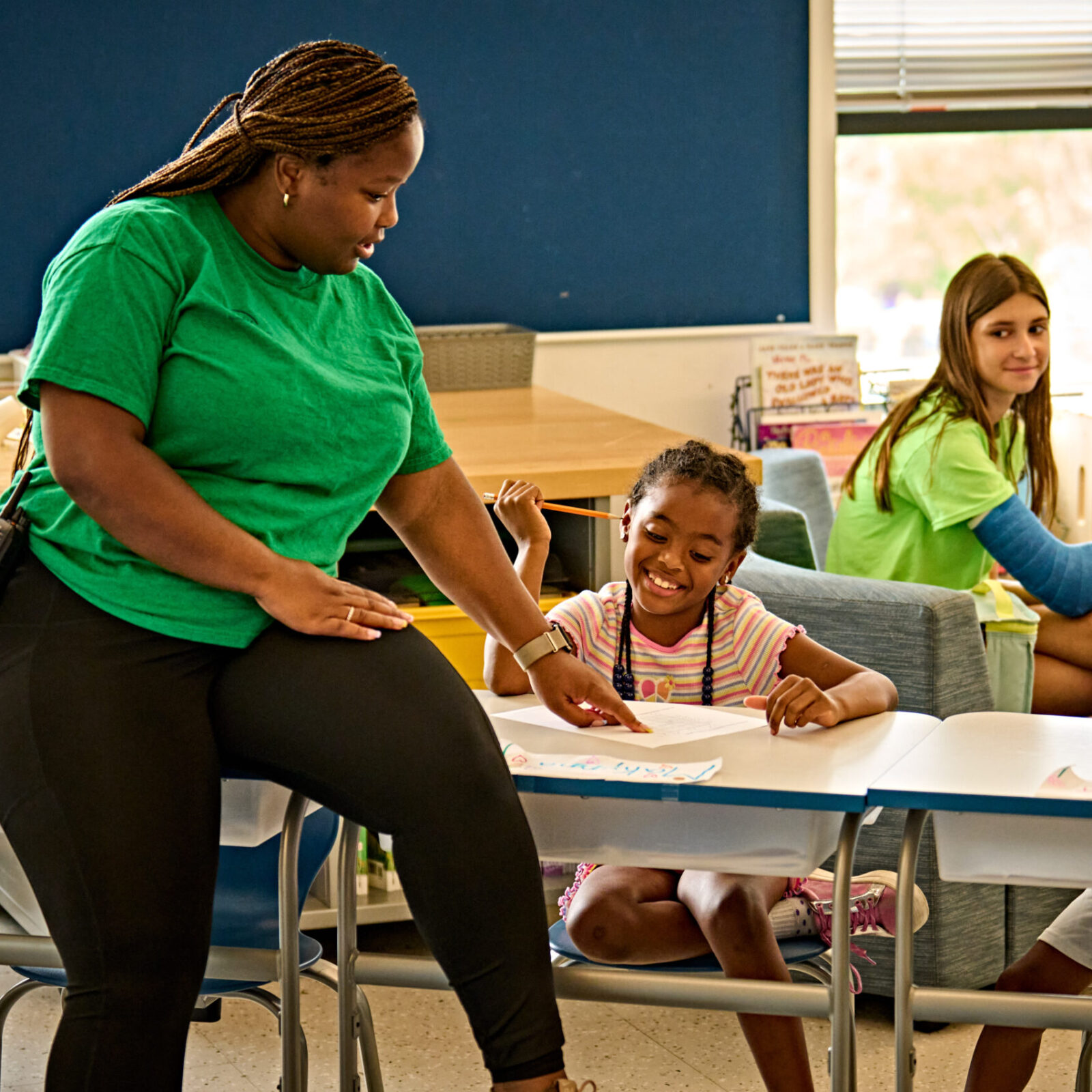 An adult wearing a green tshirt and dark pants perches on a desk in a classroom as a child smiles and looks at a paper on the desk and an older child sits in the background smiling.