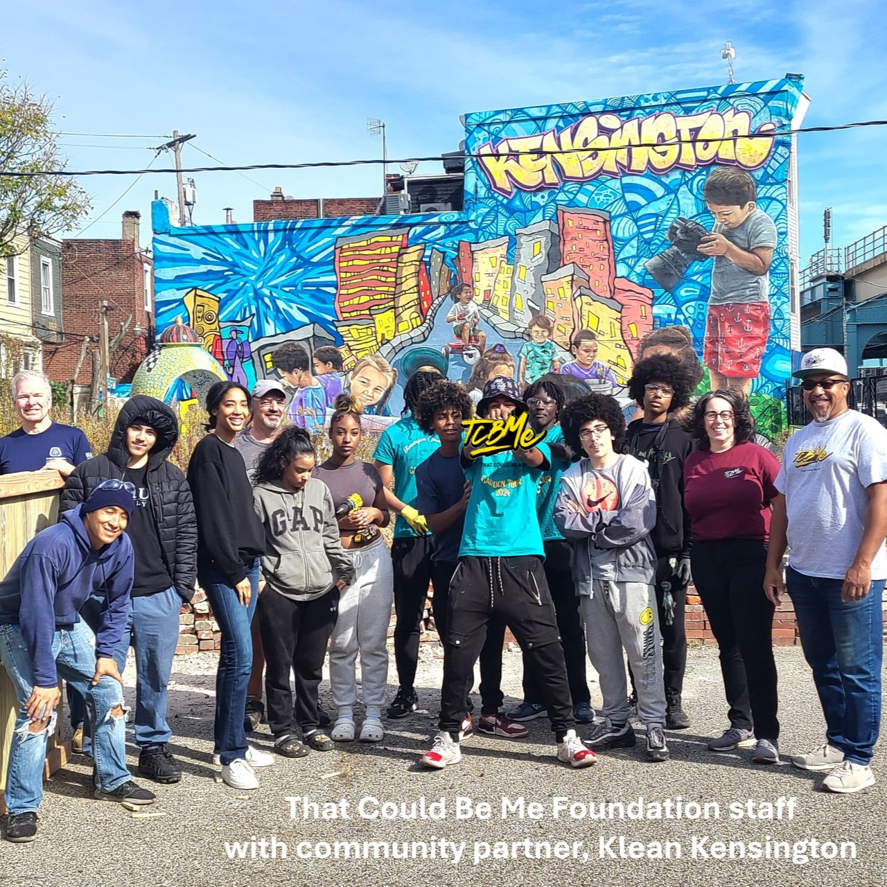 A group of adults and youth stand in a parking lot on a sunny day in front of a building painted with a multicolor mural that reads 