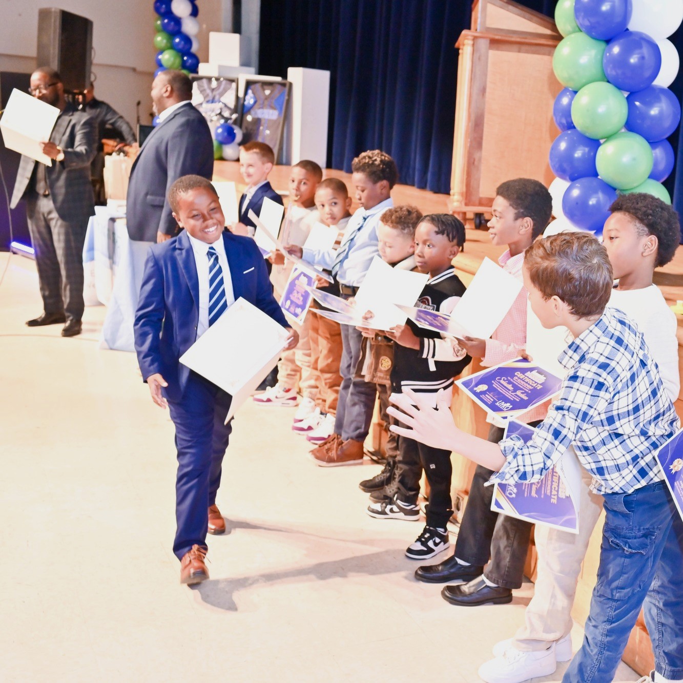A group of young boys stands on stage holding certificates during an award ceremony, with one boy in a blue suit walking confidently towards the audience, smiling. The stage is decorated with blue and green balloons, and adults are present in the background.