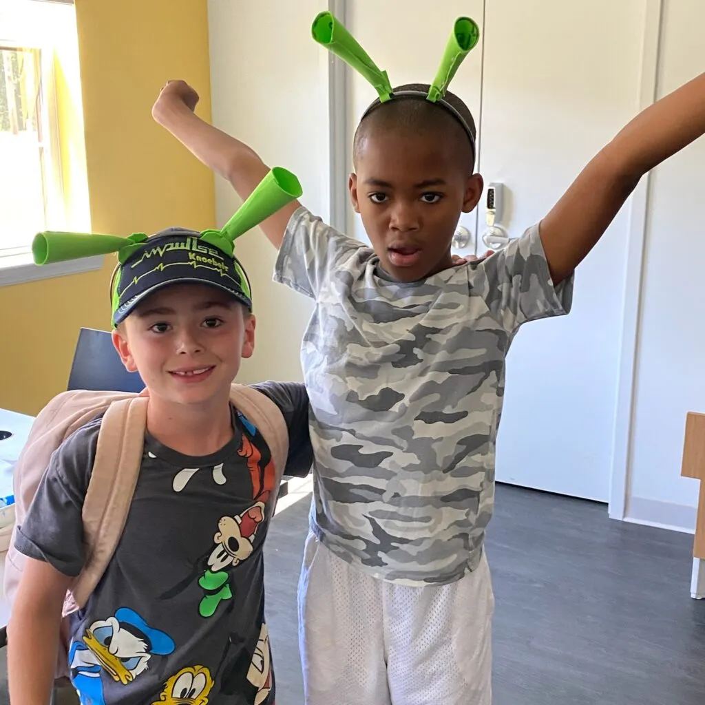 Two children are standing indoors, wearing playful green antenna headbands. The child on the left is wearing a dark t-shirt featuring cartoon characters, a baseball cap, and a backpack, smiling at the camera. The child on the right is wearing a camouflage t-shirt and white shorts, holding their arms up in an enthusiastic pose. The background shows a light-filled room with a door and a window.