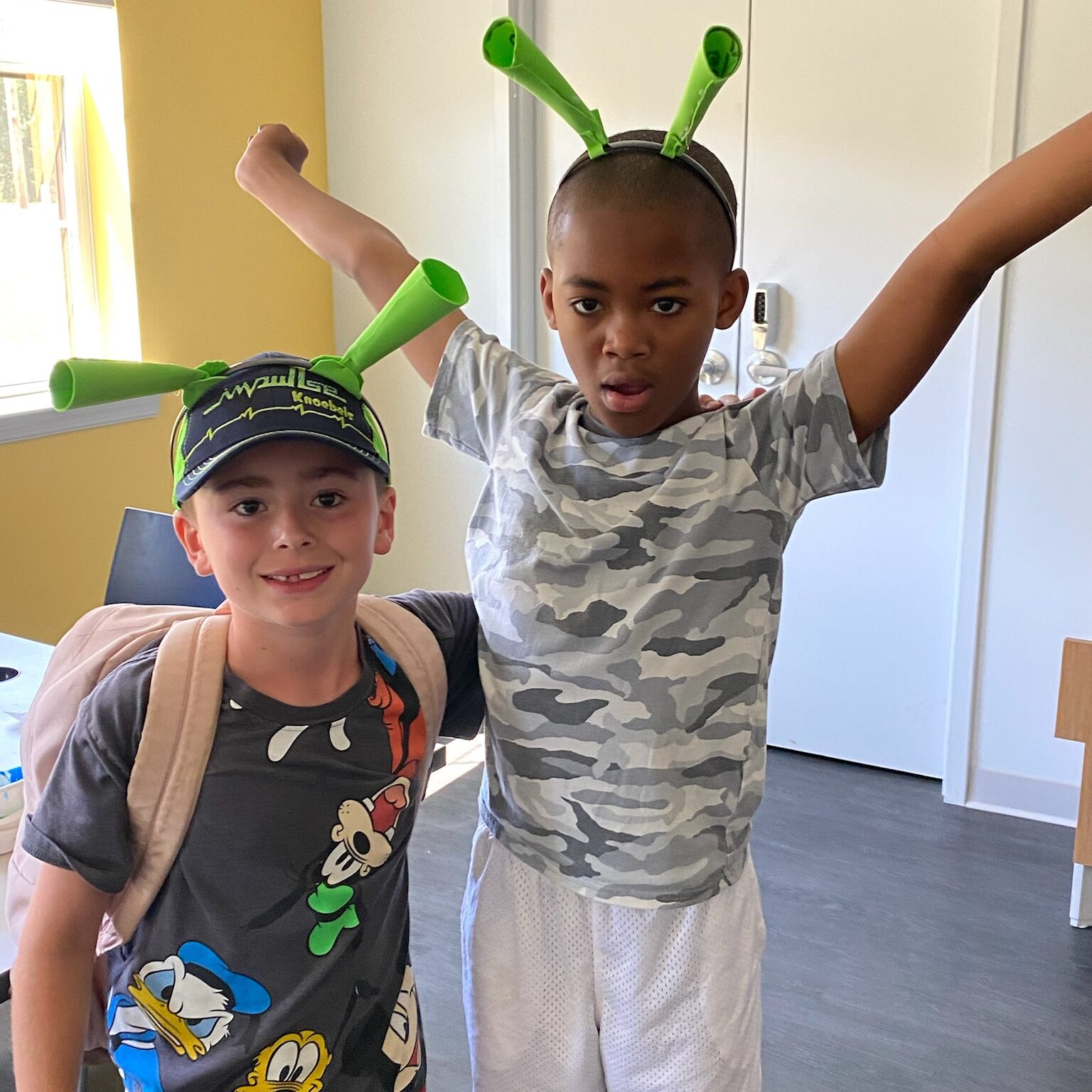 Two children are standing indoors, wearing playful green antenna headbands. The child on the left is wearing a dark t-shirt featuring cartoon characters, a baseball cap, and a backpack, smiling at the camera. The child on the right is wearing a camouflage t-shirt and white shorts, holding their arms up in an enthusiastic pose. The background shows a light-filled room with a door and a window.