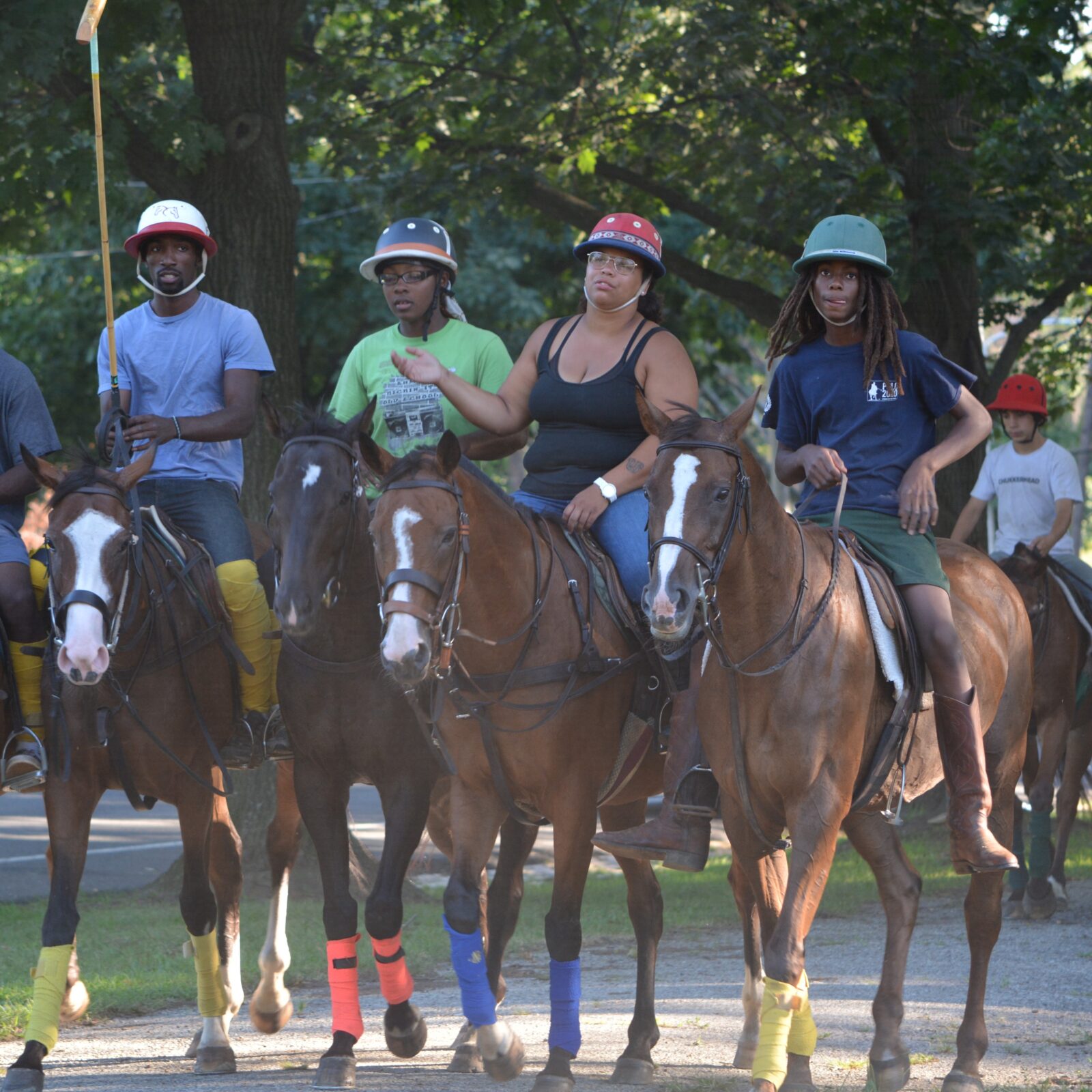 Five young people are shown riding horses in a wooded area, each wearing helmets and casual attire. The horses have colorful leg wraps, and the riders appear to be participating in an outdoor activity or training session.