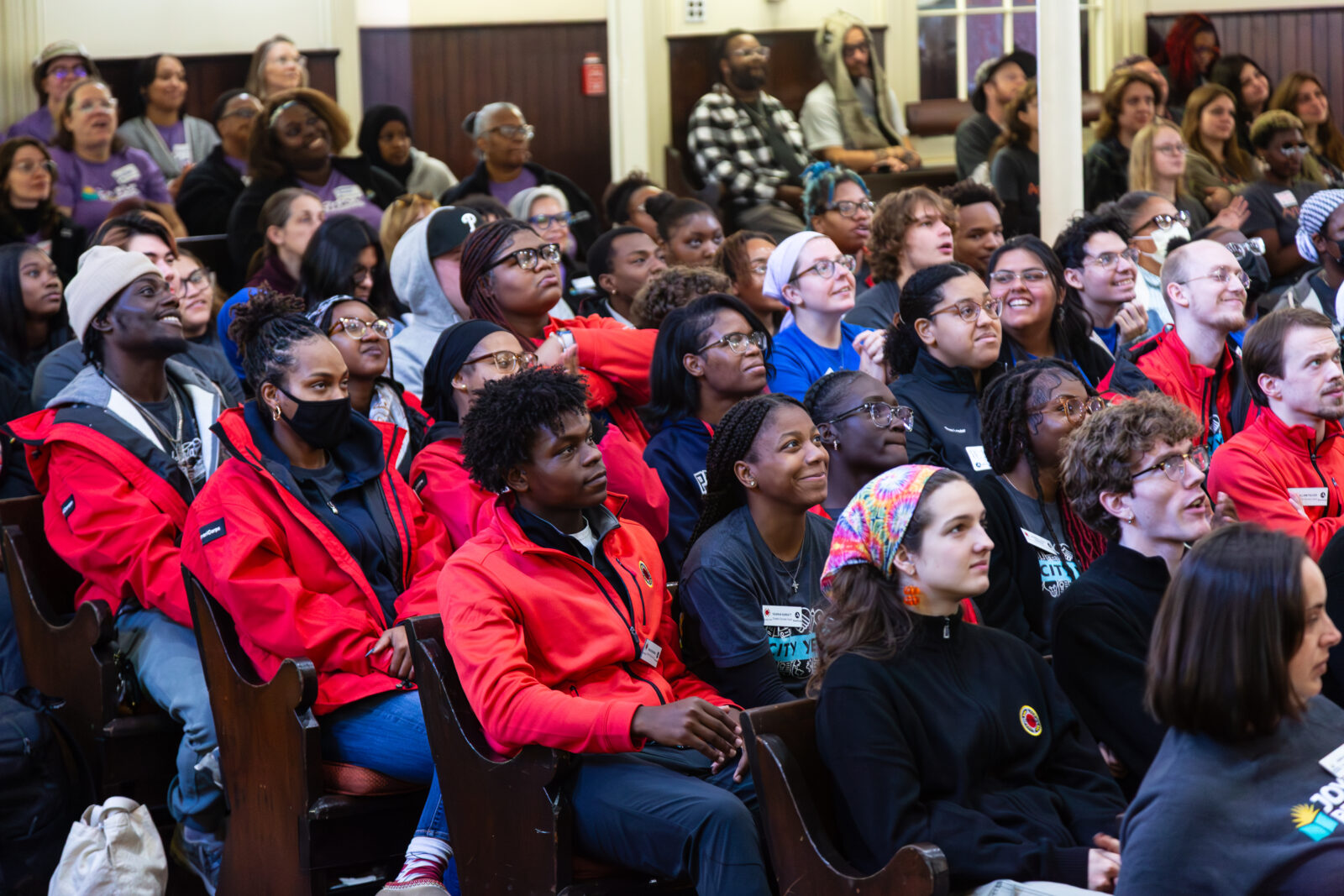 A large group of people are shown in theater-style seating, with several individuals wearing red jackets.